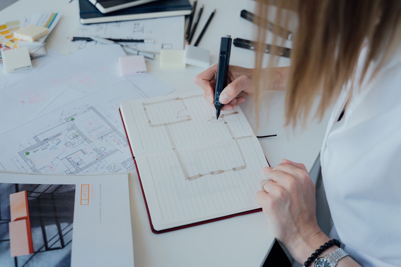 Designer sketching in a notebook beside floor plans and material swatches